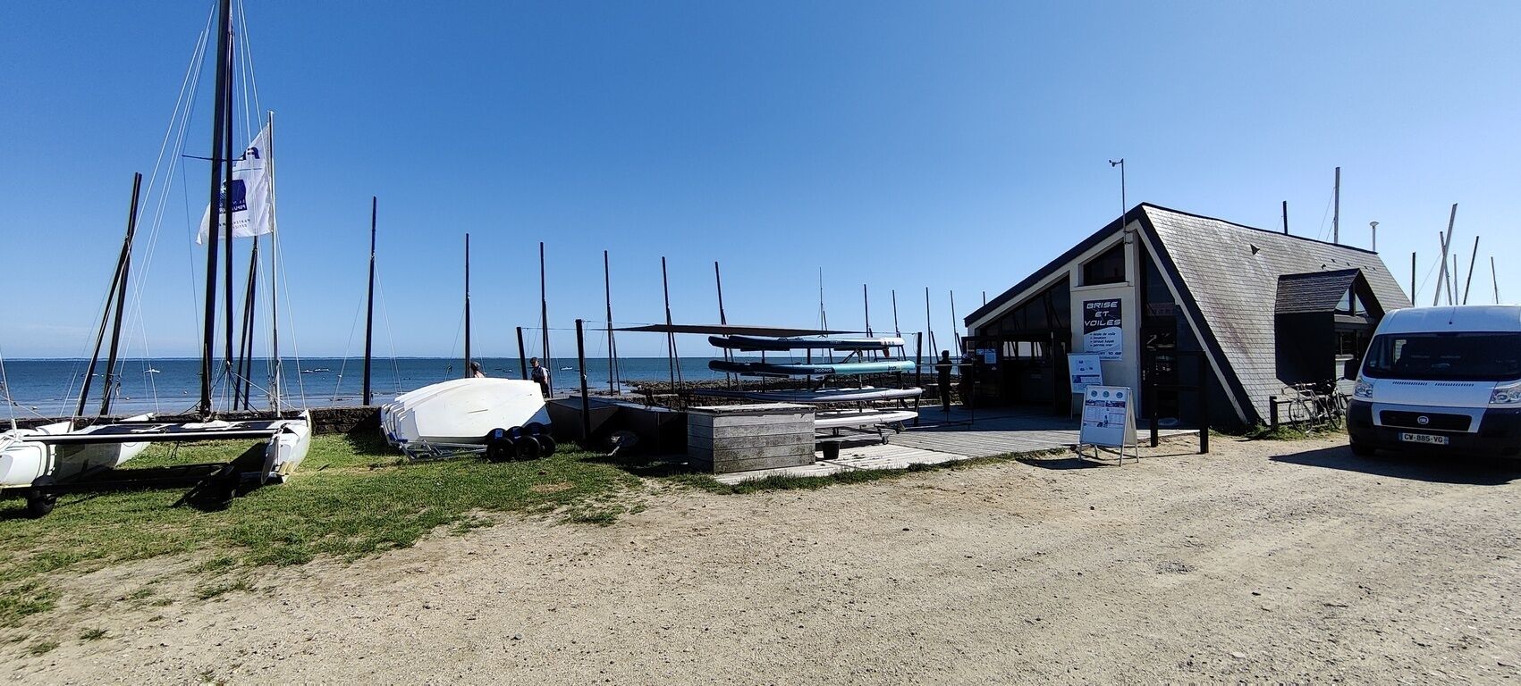 A beachside building with a sloped roof, a covered area for boats, and a parked van.