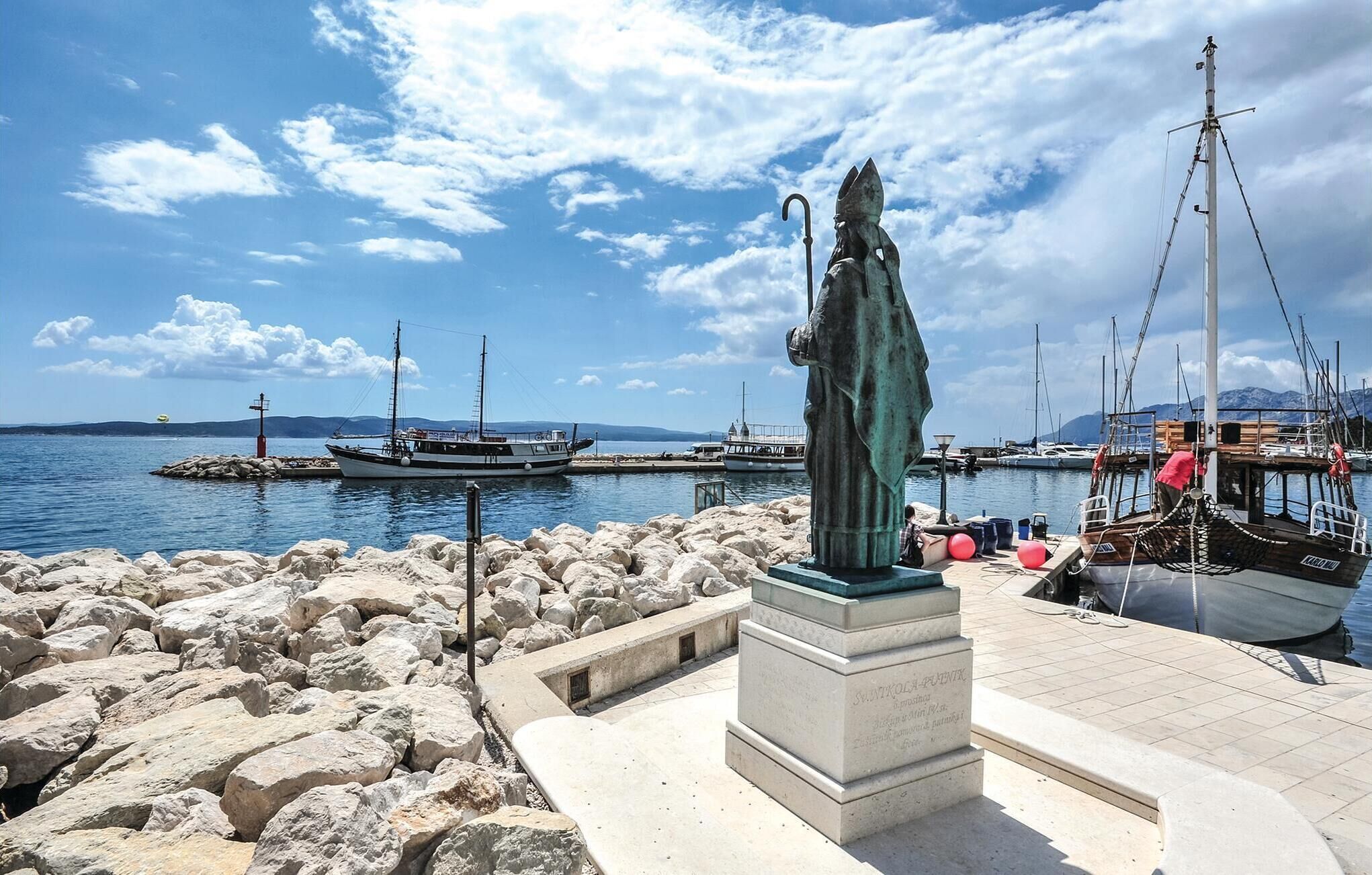 Une statue dans un port, avec des bateaux, des brise-lames rocheux et un ciel dégagé.