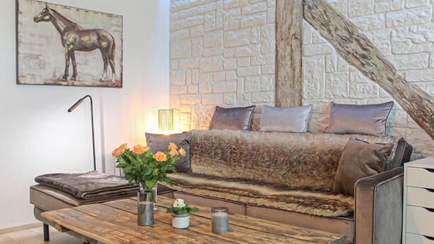 A cozy living room with a sofa, cushions, a coffee table, a vase with flowers, and a painting of a horse.