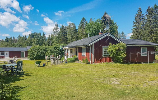 Ett rött trähus med ett grått tak, en liten veranda och en satellitmottagare. Framför huset ligger en grön gräsmatta med en skottkärra och utemöbler.