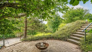 Une terrasse circulaire en briques, avec un foyer métallique au centre, entourée d’une végétation luxuriante et d’un escalier sur le côté droit.