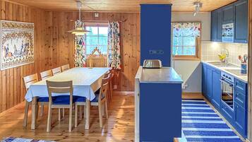 A wooden interior with a dining table, chairs, a kitchen area with blue cabinets, and a patterned rug.