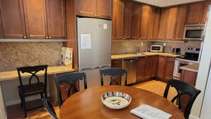 A modern kitchen with wooden cabinets, a stainless steel refrigerator, a microwave, an oven, and a dining table with chairs. - Pompano Holiday Gem (Pompano Beach)