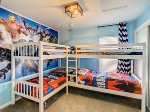 Habitación con literas, ropa de cama con temática de superhéroes, un lámpara en el techo y una ventana con persianas.