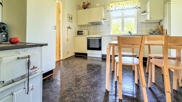 Een keuken met witte kasten, een fornuis, een magnetron, en een eethoek met houten stoelen.