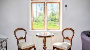 Een kamer met twee houten stoelen en een kleine ronde tafel, een groot raam met uitzicht op een tuin, en een thermostaat aan de muur.