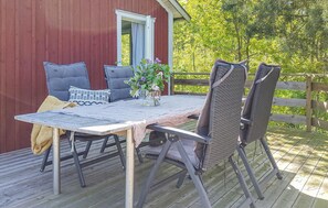 Een houten terras met een tafel, stoelen, een klein raam en uitzicht op bomen.