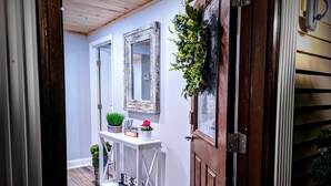 Een gang met een houten deur, een witte console tafel met planten, een spiegel en een bord met een welkomstboodschap.