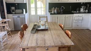 Une cuisine avec des meubles blancs, une table à manger en bois et une vue sur l’extérieur grâce à une grande fenêtre.
