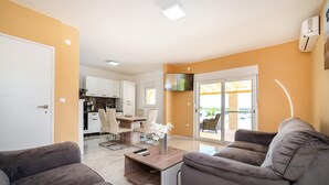 Una sala de estar moderna con un sofá, una mesa de centro y un televisor. Se ve una cocina al fondo con armarios blancos y un comedor con una mesa y sillas.