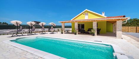 Un espace aménagé au bord de la piscine, avec un bâtiment jaune, des piliers en pierre et des sièges d’extérieur.