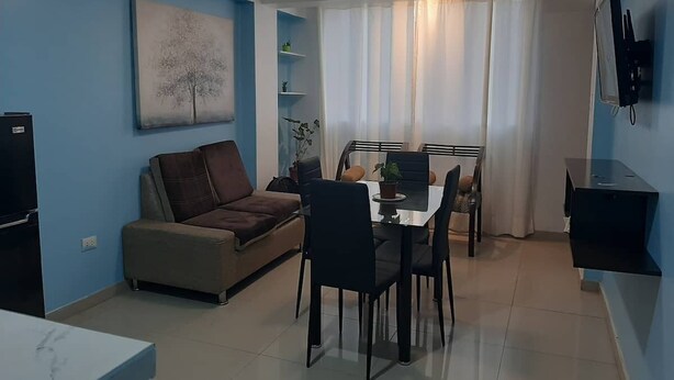 A living room with a brown sofa, a dining table with chairs, a TV mounted on the wall, and a ceiling light. - Super cozy apartment / apartment close to the airport (Callao)