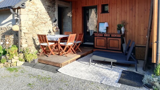 A wooden exterior with a stone wall, a covered patio with a table and chairs, and a small cabinet with a lamp and vase. - Studio Terre & Mer à Dragey (Dragey)