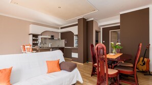 Ein modernes Wohnzimmer mit einem weißen Sofa, einem Holzespeisetisch und Stühlen. Im Hintergrund ist eine Küche mit einem Essbereich zu sehen.