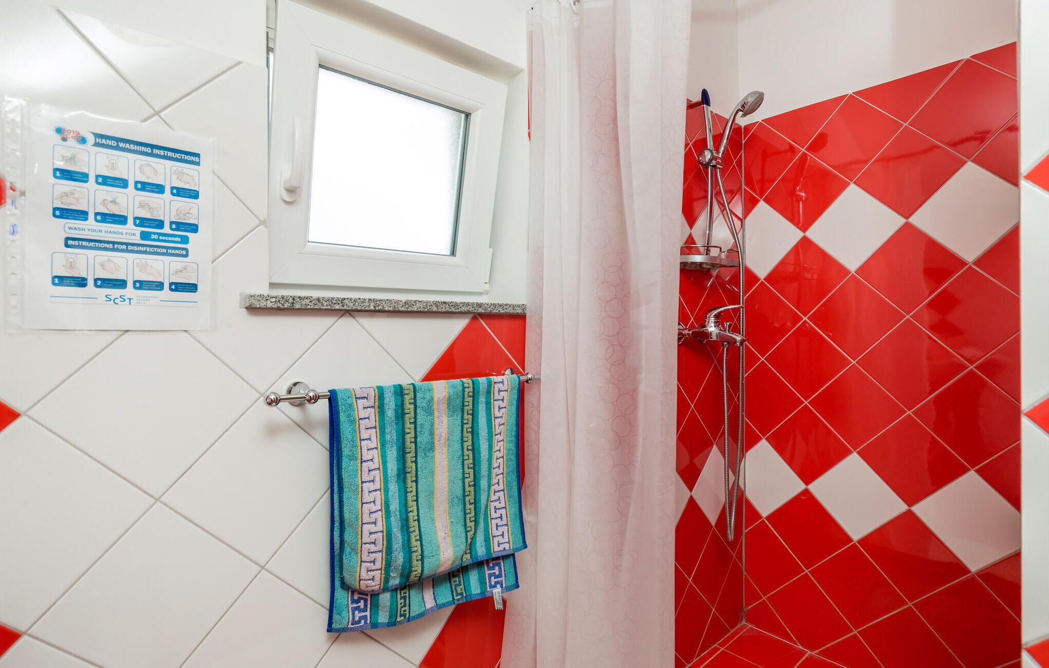 A bathroom with a red and white tiled wall, a window, a shower, and a towel rack with striped towels.