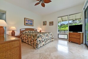 A bedroom with a bed, two bedside lamps, a ceiling fan, a TV, and a large window with a view of a green landscape.