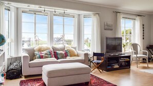 A living room with a sofa, ottoman, and a TV stand with books. Large windows provide a view of the outside. - 3 bedroom beautiful home in Vanse (Vanse)