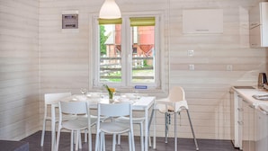 Moderni keittiö, jossa on valkoiset kaapit, ruokapöytä neljälle ja ikkuna, josta avautuu näkymä punaiseen rakennukseen ja vehreään maisemaan.