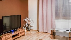 A living room with a TV on a wooden stand, a vase with white flowers, and a glass table.