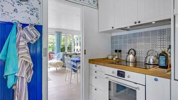 A kitchen with white cabinets, a wooden countertop, and a patterned backsplash. There is a window with curtains, a chair, and a table visible in the background.