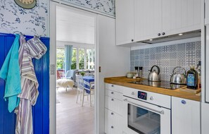 A kitchen with white cabinets, a wooden countertop, and a patterned backsplash. There is a window with curtains, a chair, and a table visible in the background.
