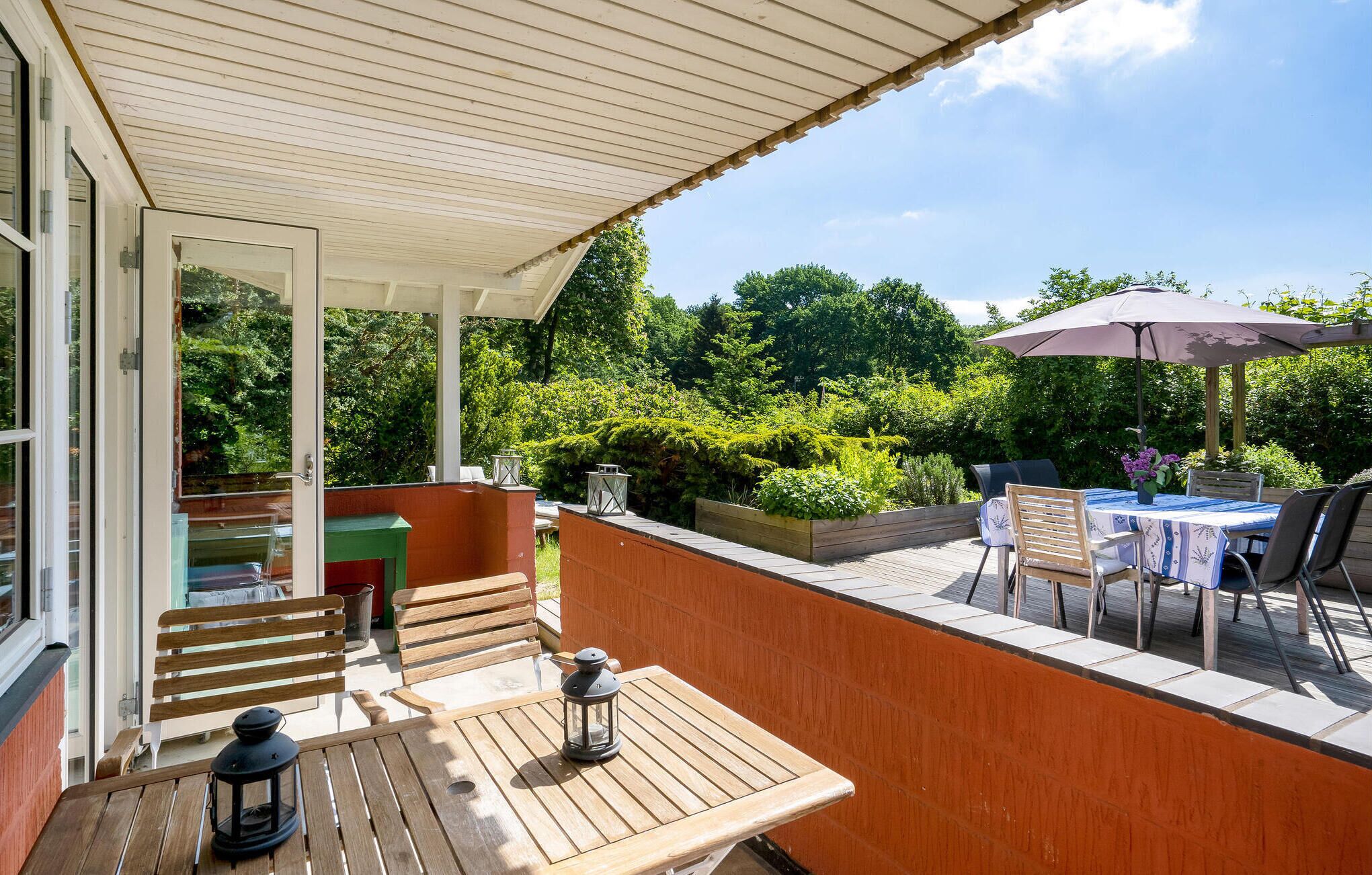 A covered outdoor patio with wooden furniture, a table with a lantern, and a view of lush greenery.