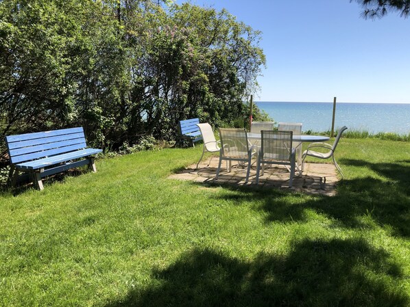 Un'area picnic con una panchina blu, sedie in metallo e un tavolo vicino a una zona erbosa con alberi e vista sull'acqua.