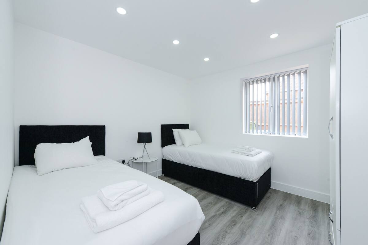 Ein modernes Hotelzimmer mit zwei Betten, einem kleinen Nachttisch mit Lampe, einem Fenster mit Rollläden und einem Kleiderschrank.