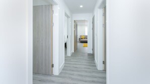 Un couloir moderne aux murs blancs et au sol gris, qui mène à une pièce contenant un canapé et un tapis jaune.
