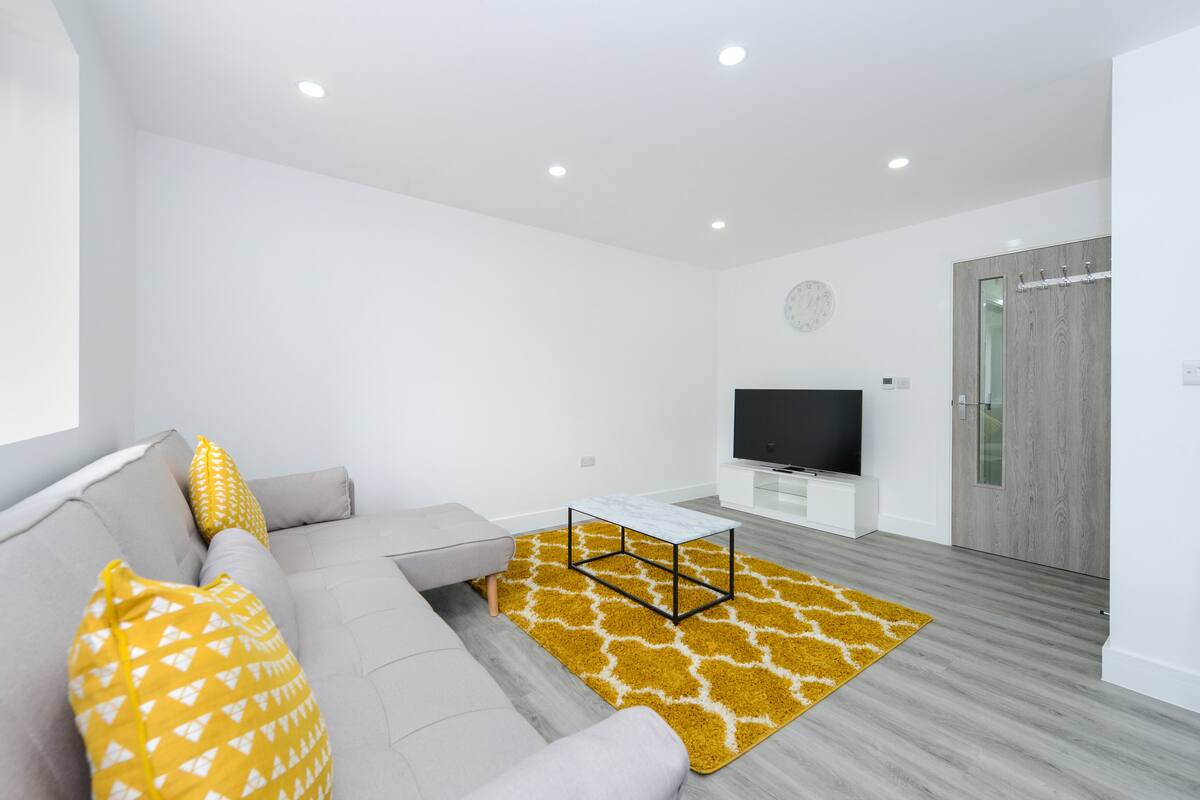Ein modernes Wohnzimmer mit einem grauen Sofa, gelben Kissen mit Muster, einem weißen Couchtisch und einem Flachbildfernseher.