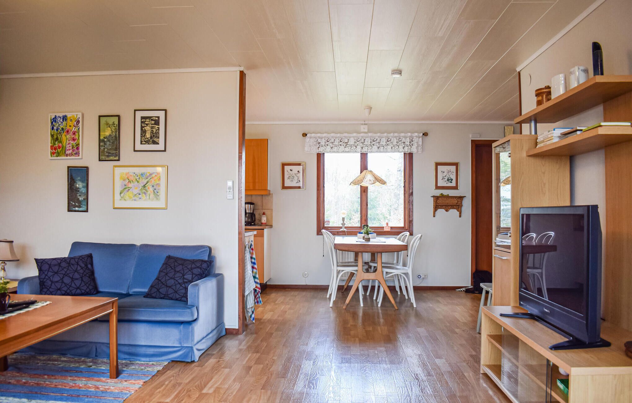 Ett vardagsrum med en blå soffa, ett träbord och en TV-bänk. Det finns en matplats med ett bord och stolar, och ett kök syns i bakgrunden.