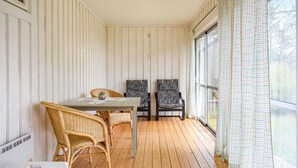 Huoneessa on puulattia, kaksi rottinkituolia, maljakko pöydällä ja ikkunassa kuviolliset verhot.