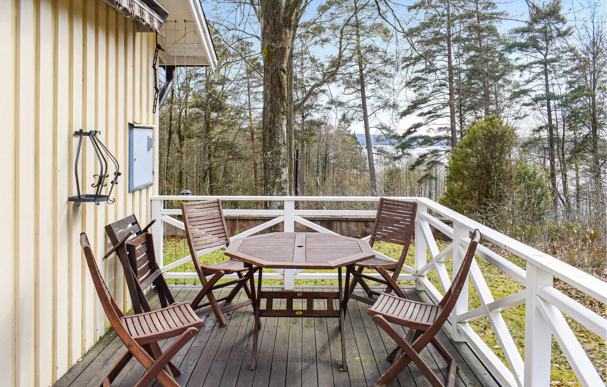 Eine Holzterrasse mit Tisch und Stühlen, umgeben von einer weißen Brüstung, mit Blick auf ein bewaldetes Gebiet.