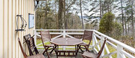 Una terraza de madera con una mesa y sillas, rodeada por una barandilla blanca, con vistas a un área boscosa.