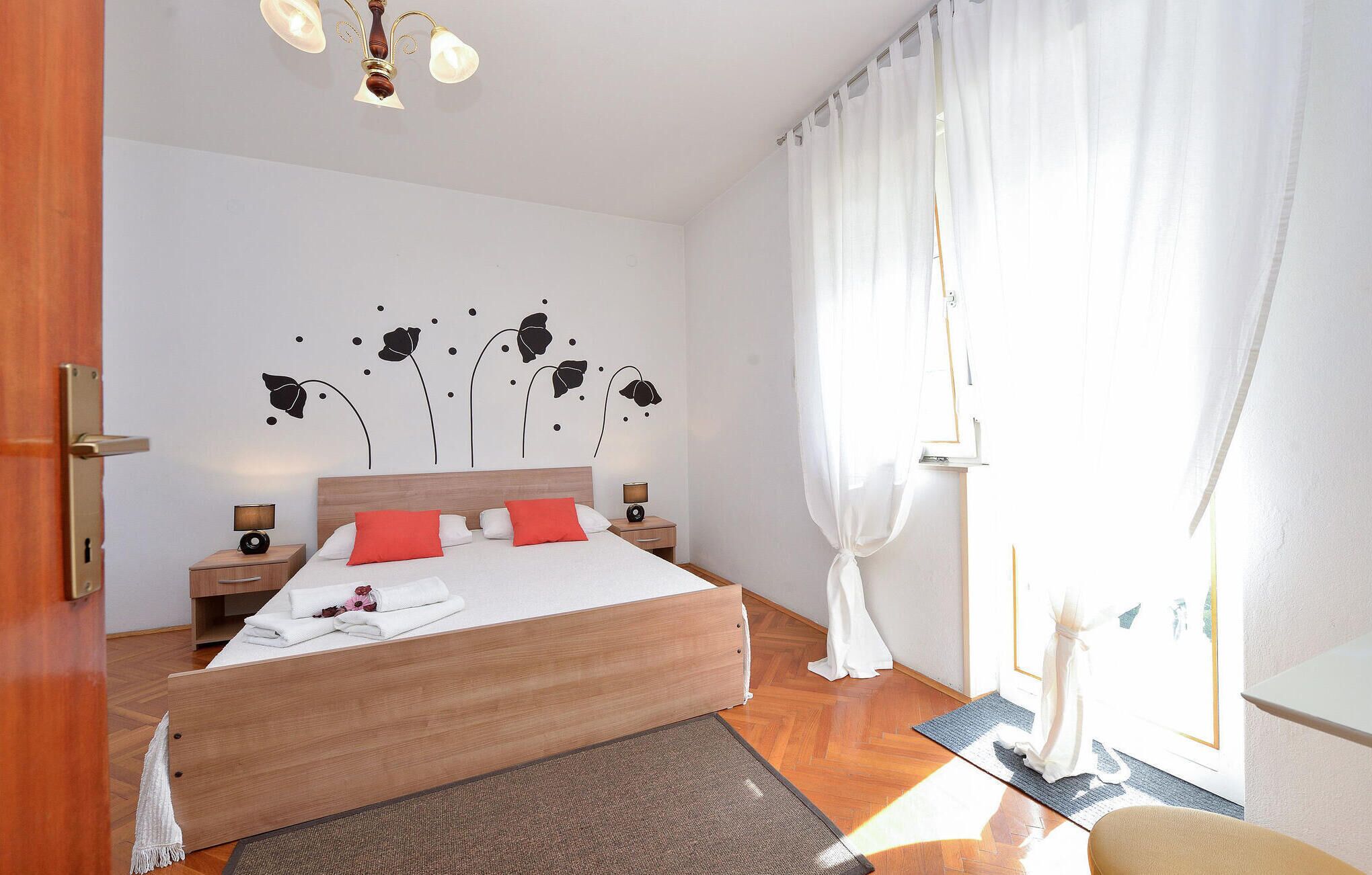 A bedroom with a wooden bed, white bedding, red pillows, a nightstand with a lamp, a wall-mounted light fixture, and sheer curtains.