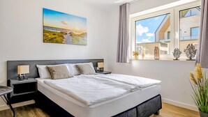 Quarto moderno com cama grande, duas mesas de cabeceira com abajures, janela com cortinas e pintura de paisagem costeira na parede.