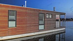 A modern floating house with horizontal wooden slats, large windows, and a metal railing overlooking calm water.