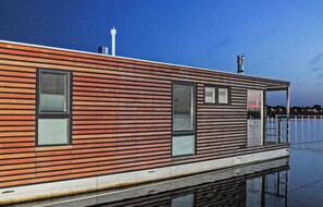 Une maison flottante moderne, dotée de lambris en bois horizontaux, de grandes fenêtres et d’un garde-corps en métal surplombant une étendue d’eau calme.