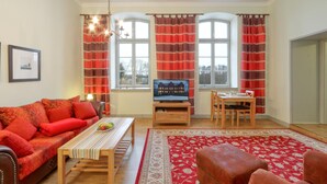 A living room with a sofa, a coffee table with fruit, a TV on a wooden stand, a dining table with chairs, and a red patterned rug. - 1 bedroom awesome apartment in Krakow am See (Krakow am See)