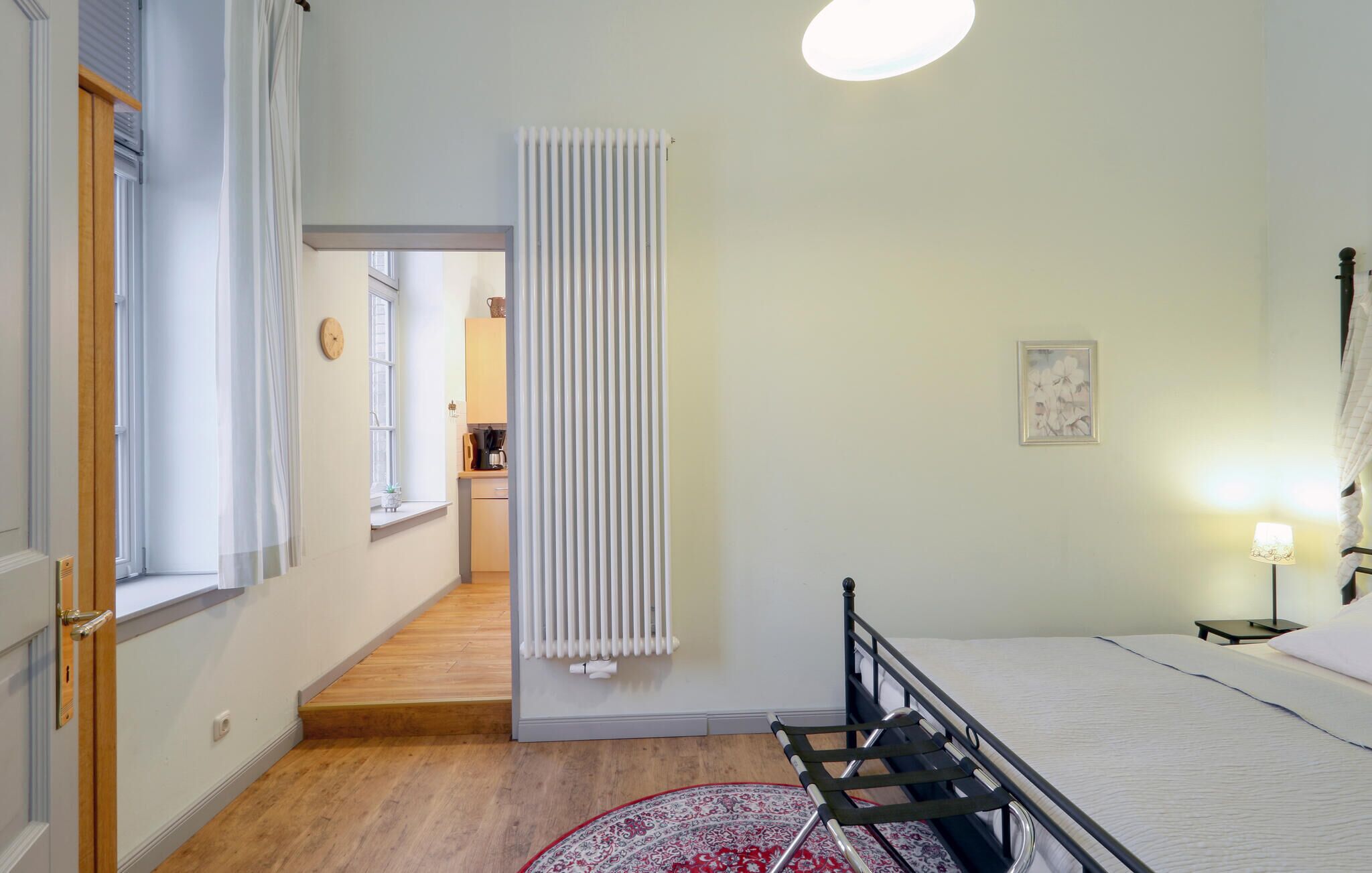A bedroom with a bed, a radiator, a window, a door, and a hallway leading to a kitchen.