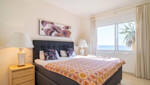 Un dormitorio con una cama, dos mesitas de noche, un velador, una ventana con cortinas y un cuadro en la pared.