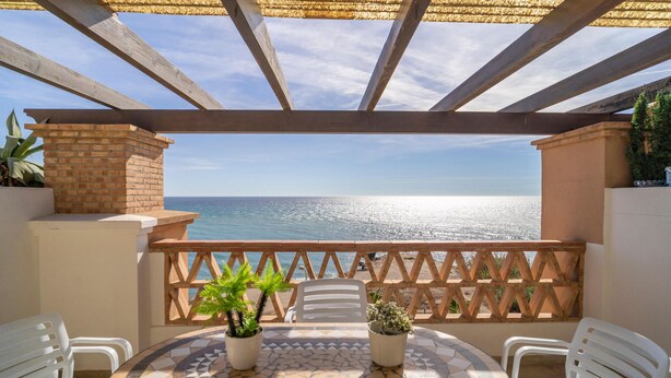Una terraza con vista al mar, que incluye una mesa de baldosas, plantas en macetas y sillas de plástico blanco.