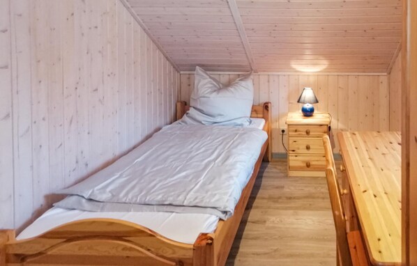 A small, single-bed room with wooden walls and a sloped ceiling. There is a bedside table with a lamp and a drawer, and a bench against the wall.