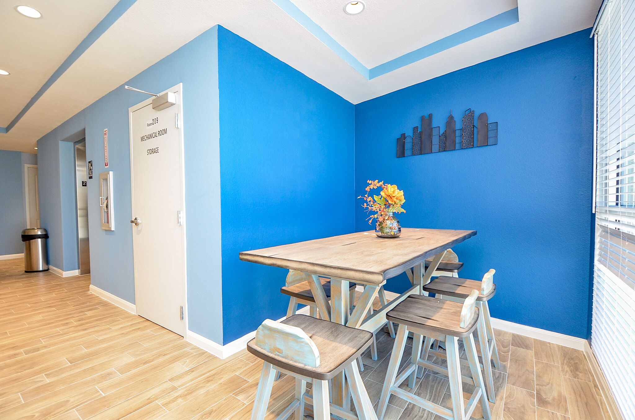 Ein Zimmer mit blauer Wand, einem Holztisch mit vier Stühlen, einer Vase mit Blumen und einer Tür mit der Aufschrift 'Mechanical Room Storage'.