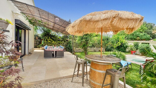 Um pátio com mesa e cadeiras, um guarda-sol de palha, um barril e uma piscina.