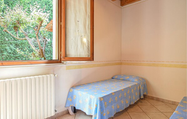 A small, single-bed room with a window overlooking greenery, a radiator, and a simple blue tablecloth.