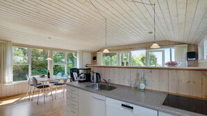 Una cocina moderna con armarios de madera, encimera de granito y lavavajillas integrado. Hay dos lámparas colgantes, una ventana con vista a zonas verdes y una zona de comedor con mesa y sillas.