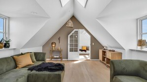 Una habitación estilo loft con techo inclinado, un sofá verde, una mesita con lámpara y un armario de madera.
