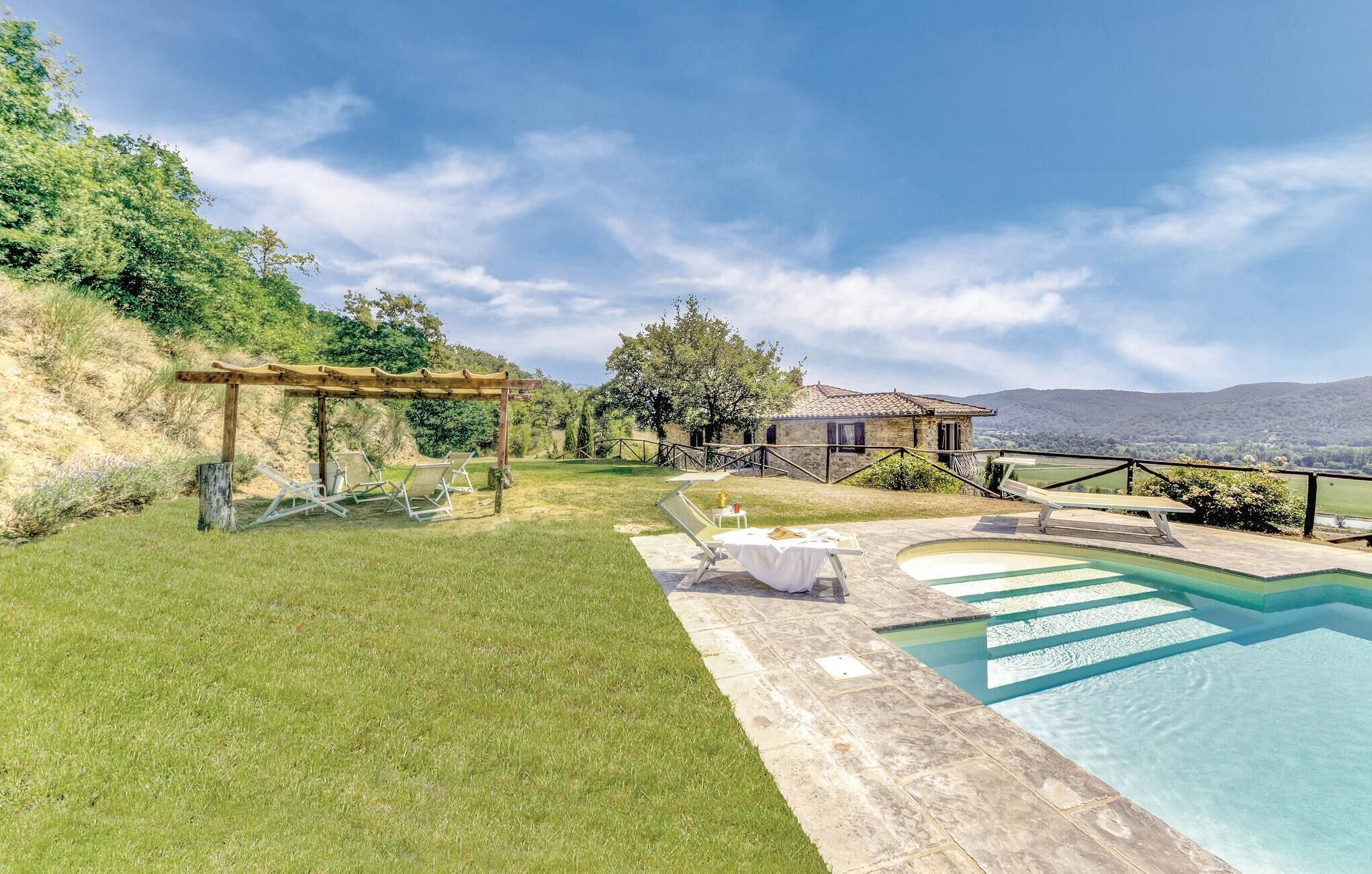 A pool area with lounge chairs, a pergola, and a stone building with a view of the hills.