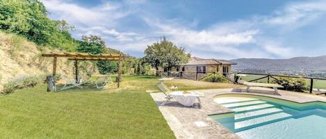 A pool area with lounge chairs, a pergola, and a stone building with a view of the hills.